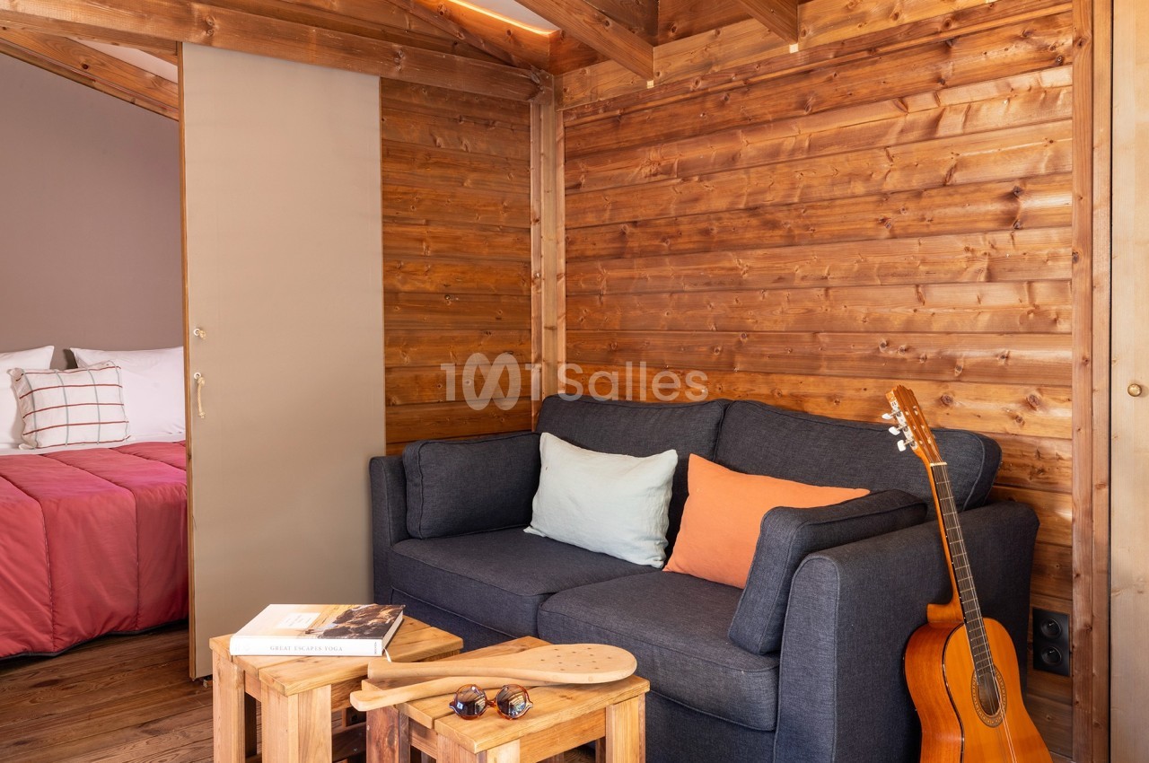 Salon en bois avec canapé gris, coussins colorés, guitare posée à côté et chambre visible en arrière-plan.