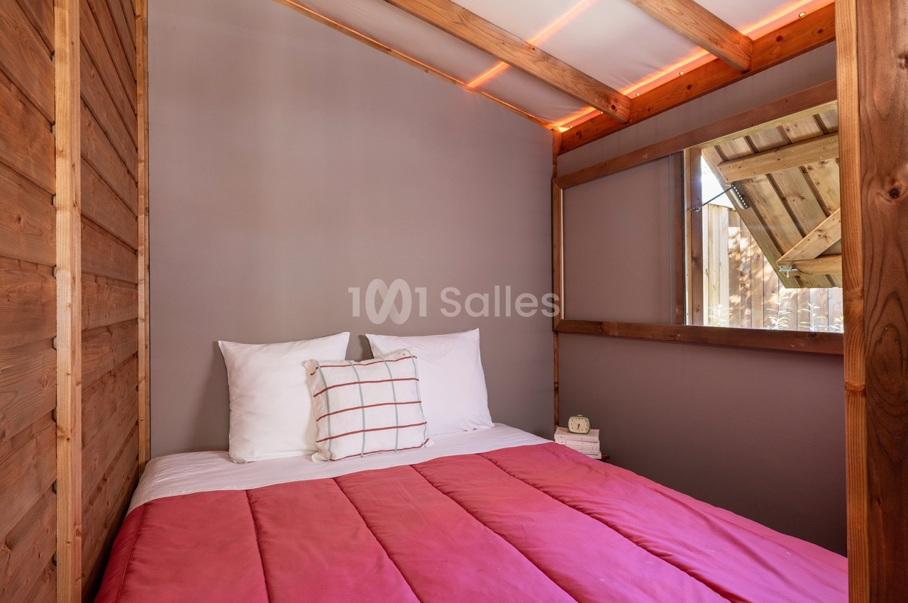 Lit double avec couette rose et oreillers blancs dans une chambre en bois avec une fenêtre donnant sur l'extérieur.