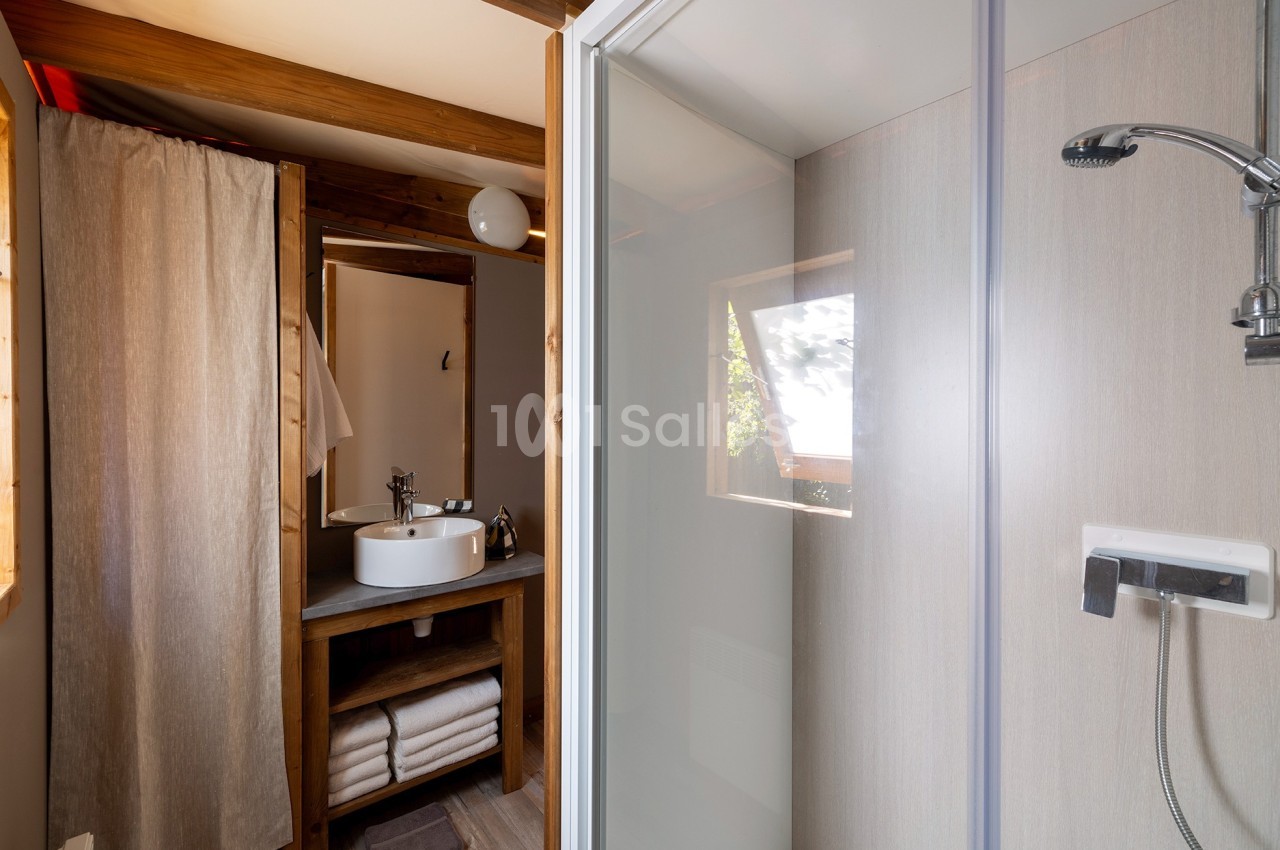 Salle de bain avec douche vitrée, lavabo sur meuble en bois, miroir, étagères avec serviettes et fenêtre lumineuse.
