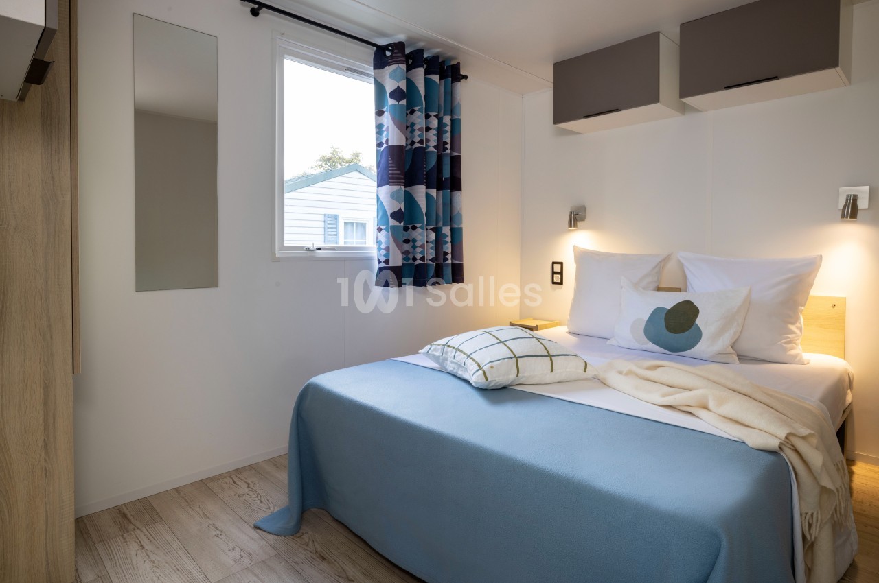 Chambre lumineuse avec lit double, draps bleus, coussins décoratifs, fenêtre avec rideaux géométriques et éclairage mural.