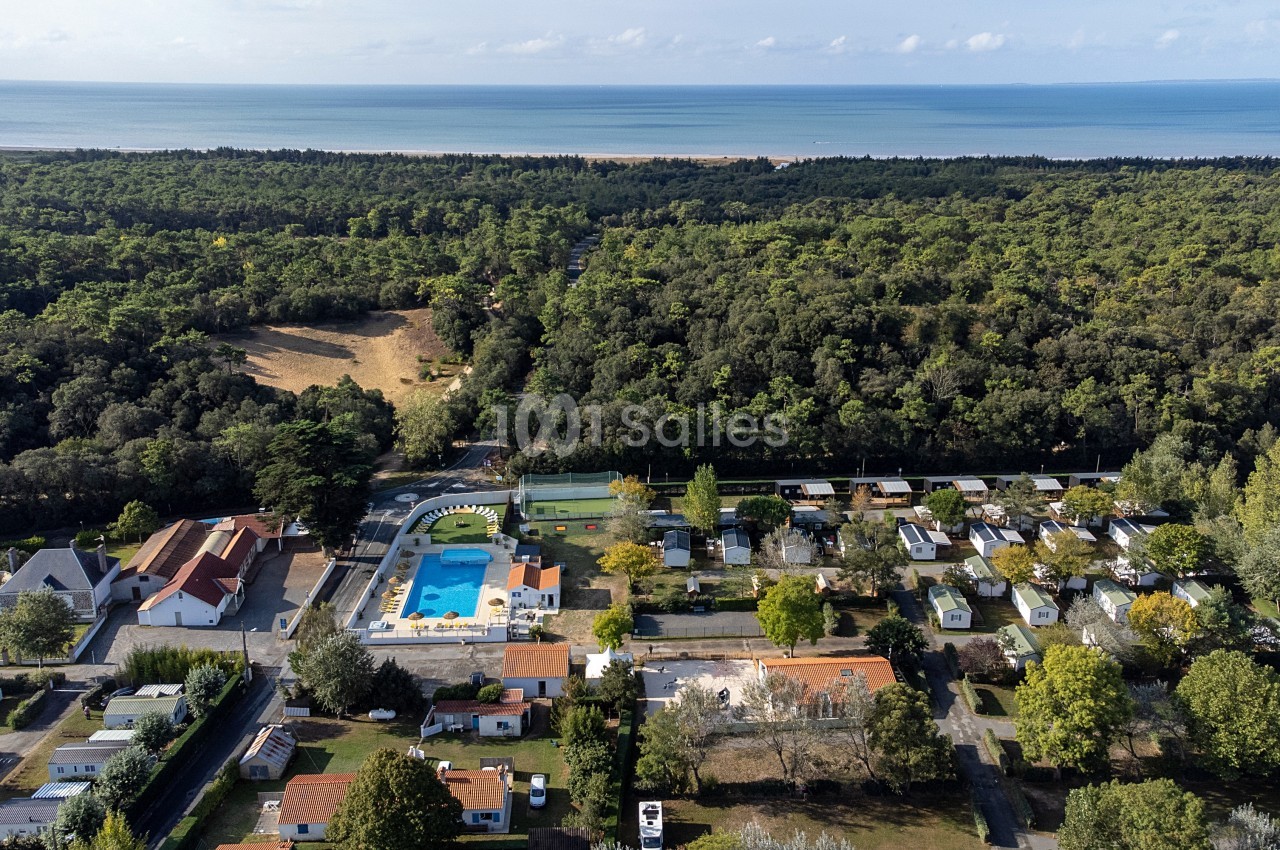 Vue aérienne d'un camping avec piscine, mobil-homes et arbres, situé près d'une forêt et d'une plage en arrière-plan.