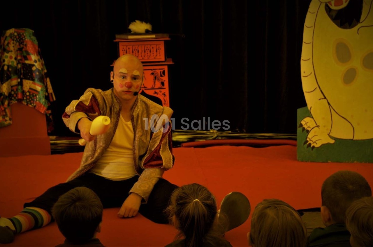 Un clown assis sur une scène rouge montre une banane à un groupe d'enfants attentifs.