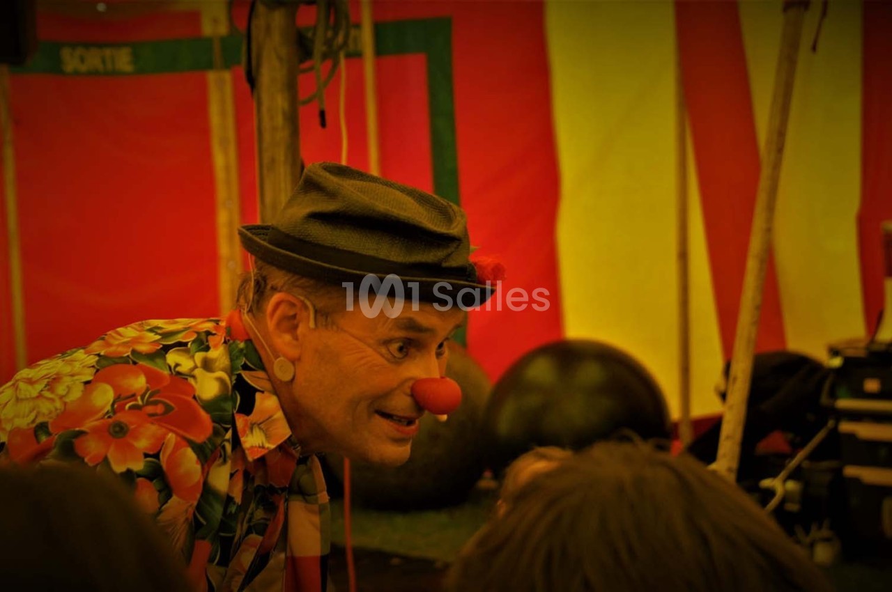 Un clown portant un chapeau et une chemise colorée interagit avec des enfants sous un chapiteau.