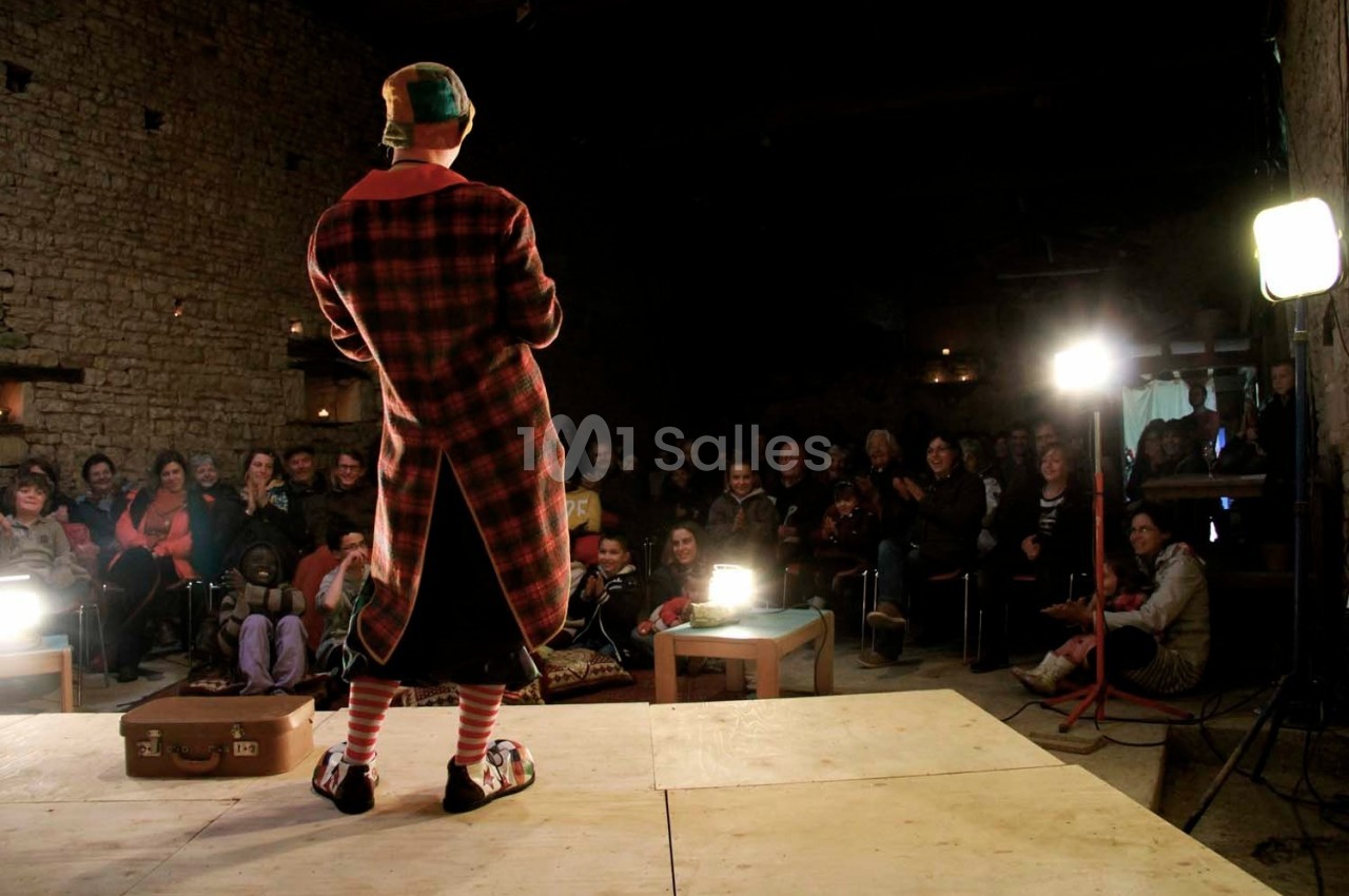 Un artiste en costume coloré se produit sur scène devant un public assis dans une salle en pierre éclairée.