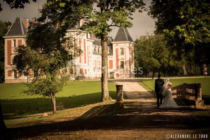 Un couple de mariés marche sur une allée bordée d'arbres, menant à un château entouré de verdure.