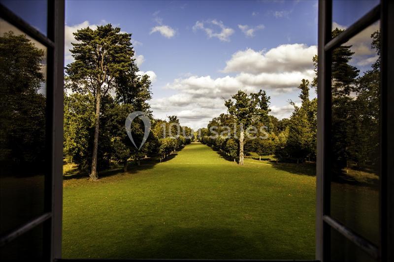 Vue depuis une fenêtre ouverte sur une vaste pelouse bordée d'arbres sous un ciel partiellement nuageux.