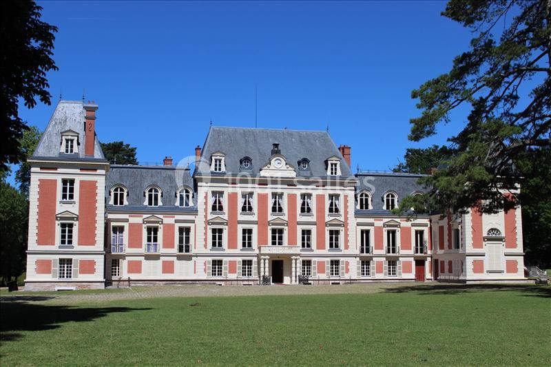 Façade d'un château avec des murs rouges et blancs, entouré de pelouse et d'arbres sous un ciel bleu.