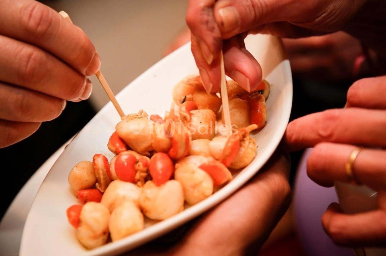 Des mains tiennent une assiette de brochettes apéritives composées de crevettes et tomates cerises.