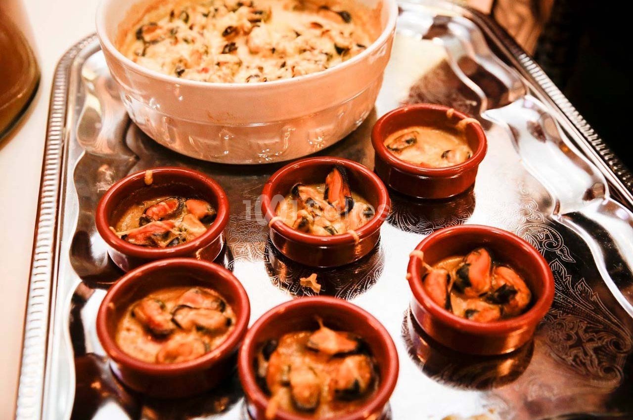 Des ramequins de moules gratinées disposés sur un plateau argenté, avec un plat principal en arrière-plan.