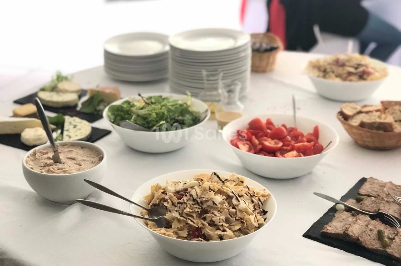 Table dressée avec des plats variés : salades, fromages, pain, pâté et assiettes empilées en arrière-plan.