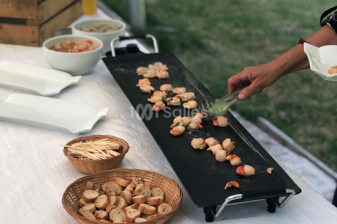 Personne cuisant des fruits de mer sur une plancha, avec des paniers de pain et des assiettes vides à proximité.