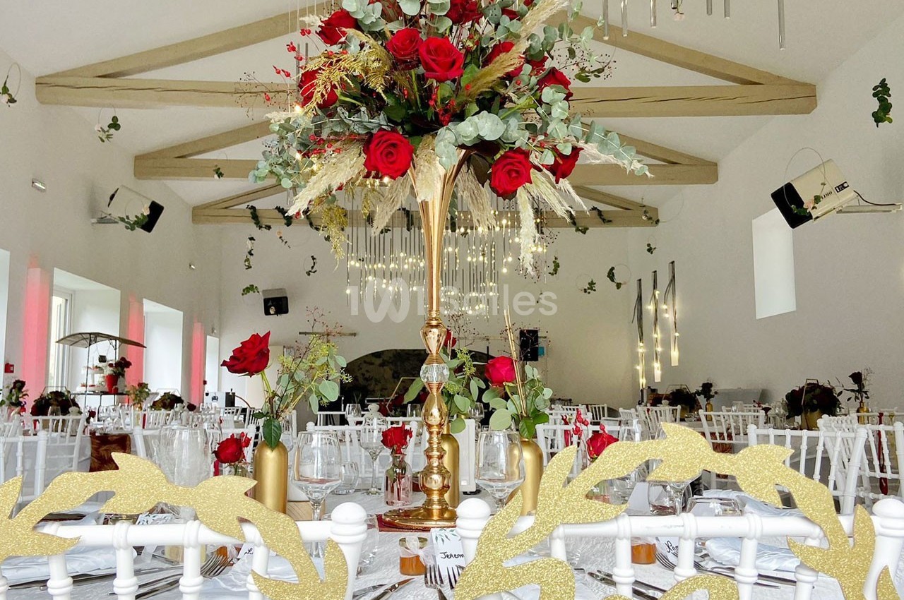 Centre de table floral avec roses rouges et feuillage, dans une salle de réception décorée avec des lumières suspendues.
