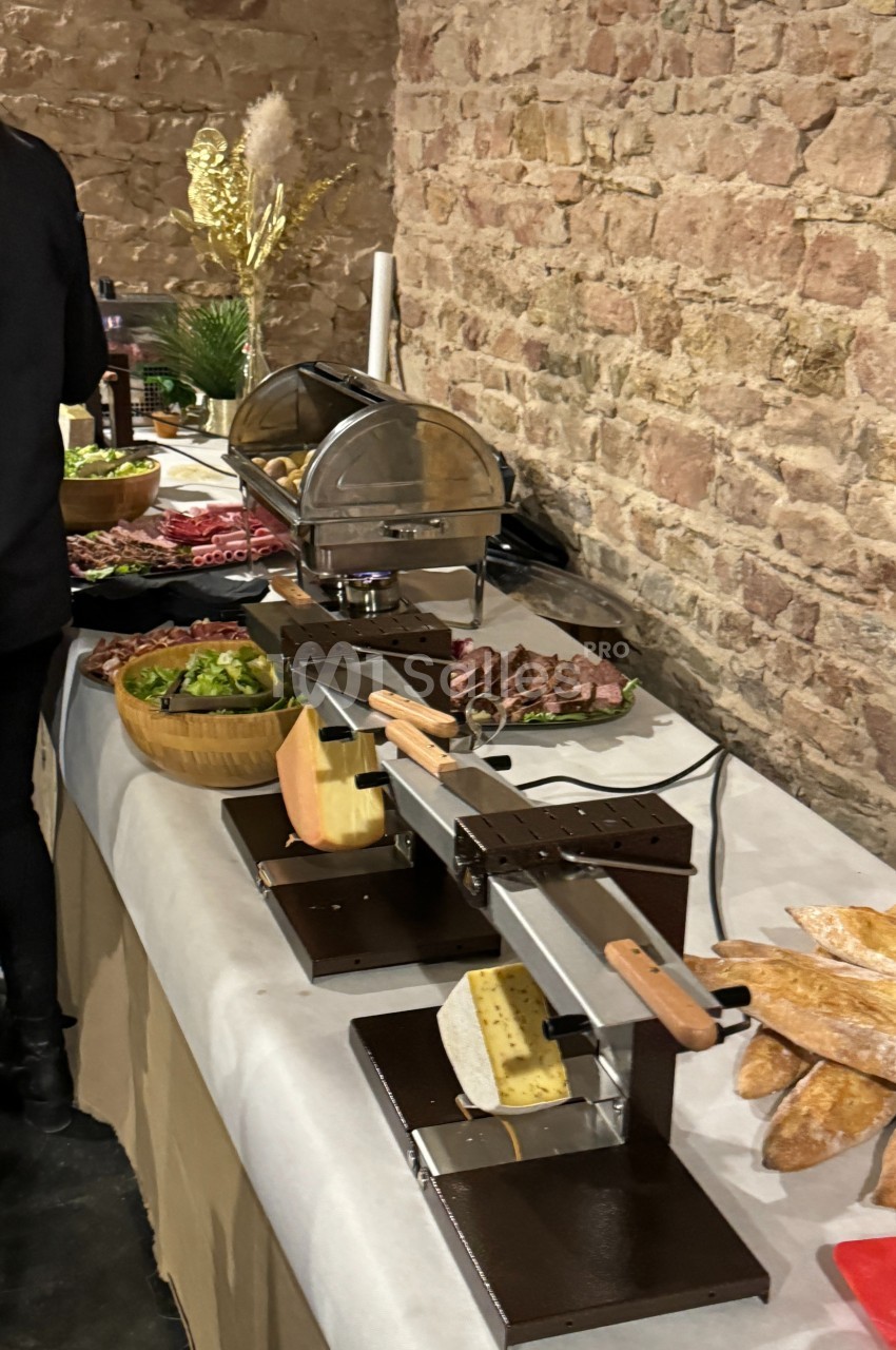 Table avec appareils à raclette, charcuterie, pain, légumes et accompagnements, dans un cadre en pierre rustique.