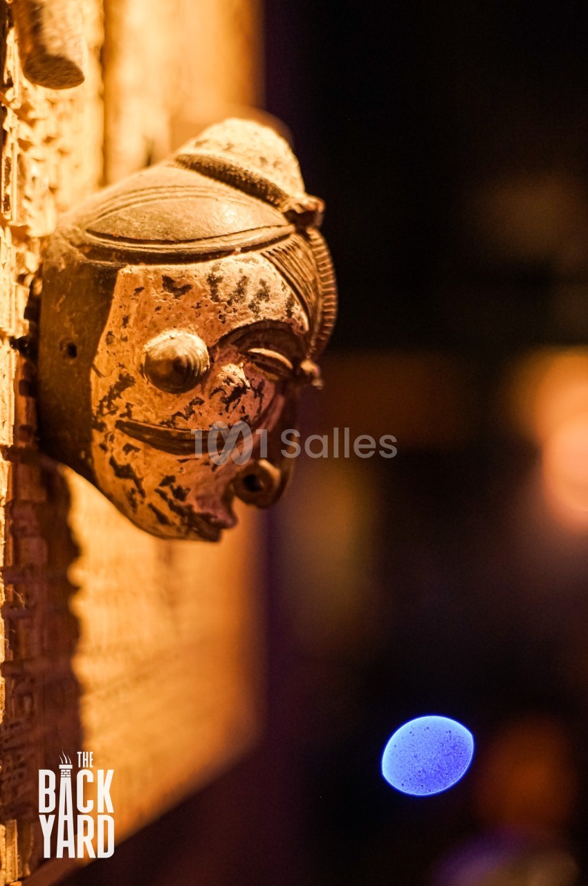 Masque sculpté en bois accroché à un mur, éclairé par une lumière chaude, avec un arrière-plan flou.