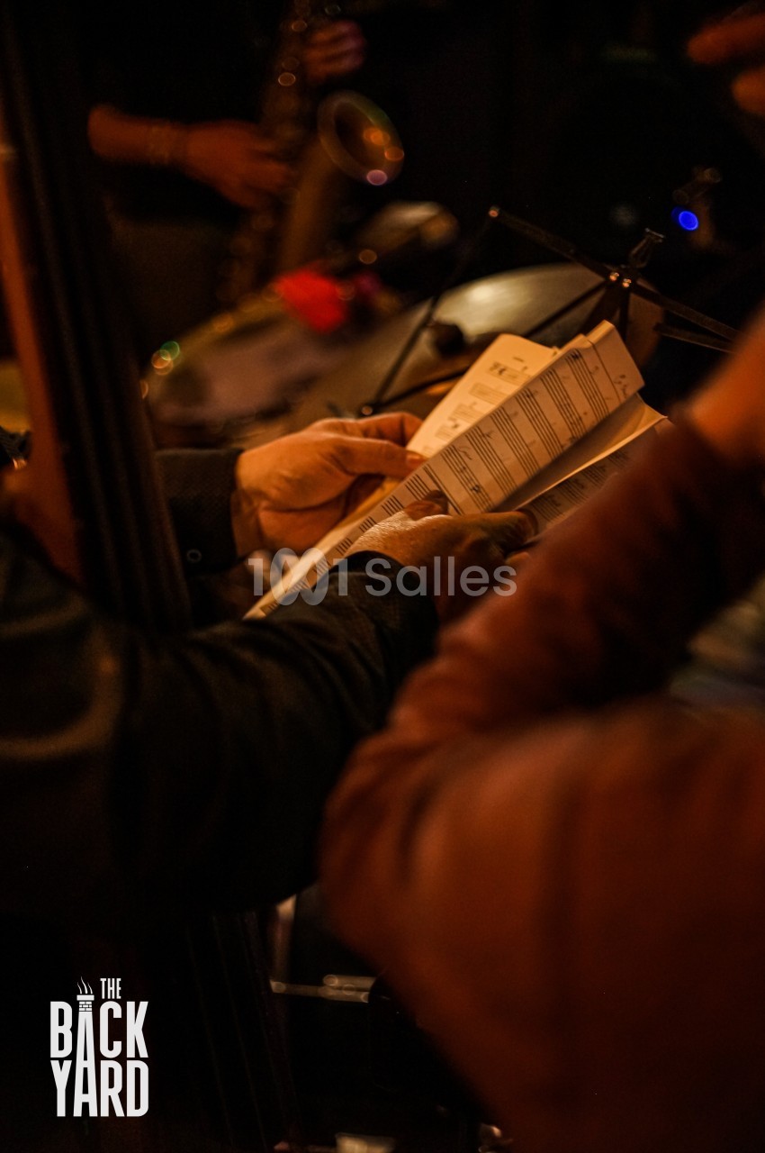 Des musiciens lisent une partition dans une ambiance tamisée, avec un saxophone visible en arrière-plan.