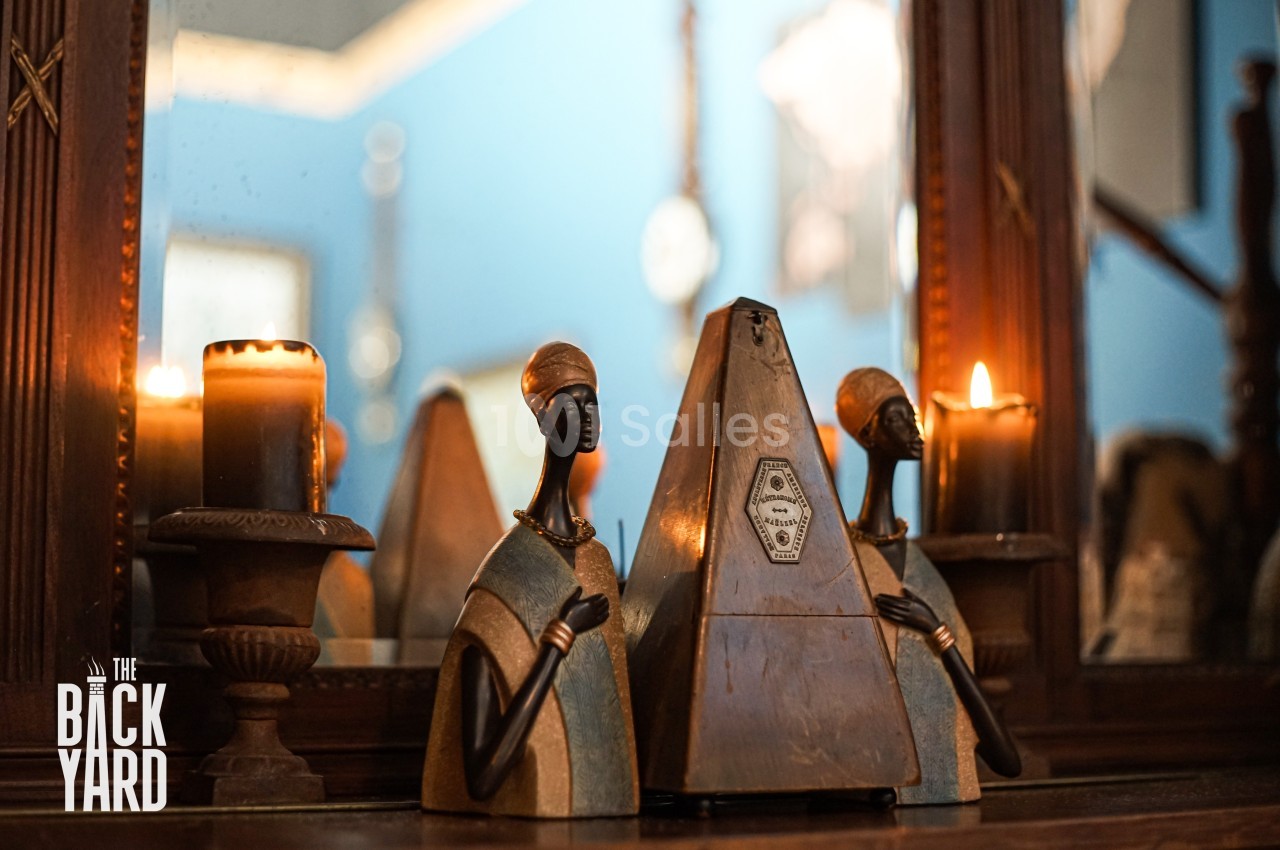 Deux statuettes africaines encadrent un métronome en bois, posés sur une table devant un miroir et des bougies allumées.