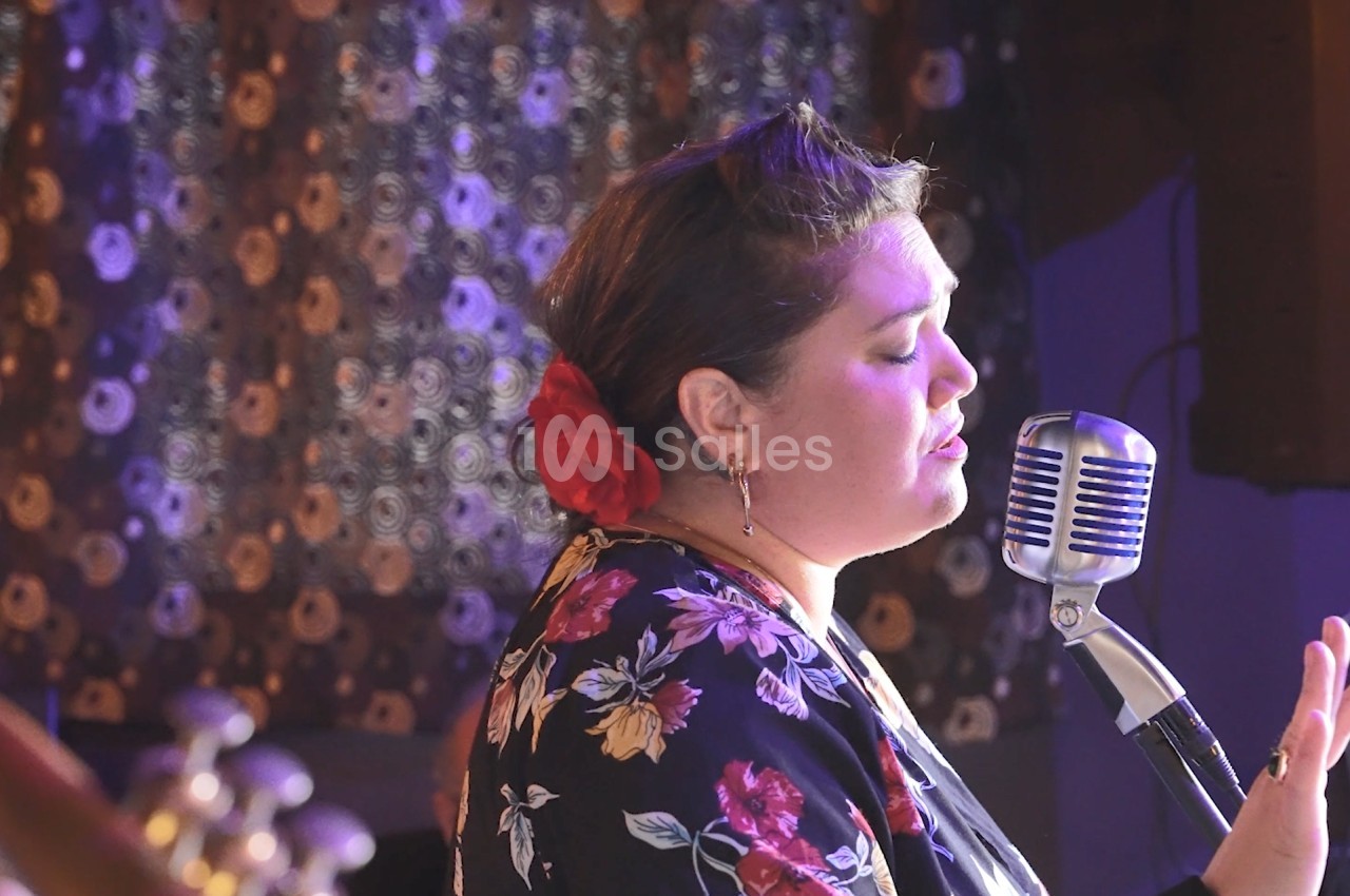 Une femme chante au micro dans une ambiance rétro, portant une robe à motifs floraux et une fleur rouge dans les cheveux.