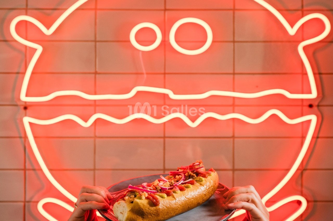 Un hot-dog garni est tenu devant un néon rouge en forme de burger sur un mur carrelé.