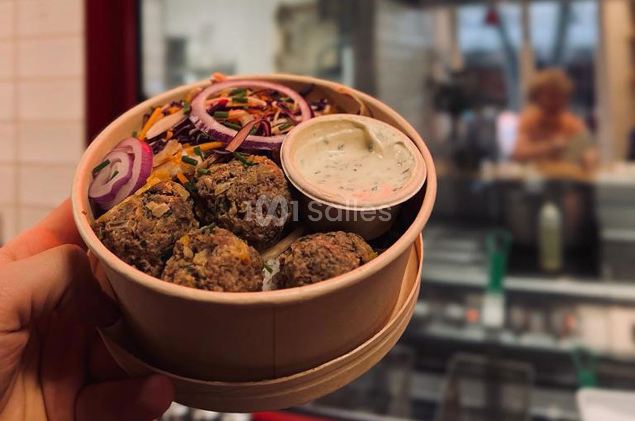 Bol en carton contenant des boulettes végétariennes, crudités colorées et une sauce blanche, tenu devant une cuisine ouverte.