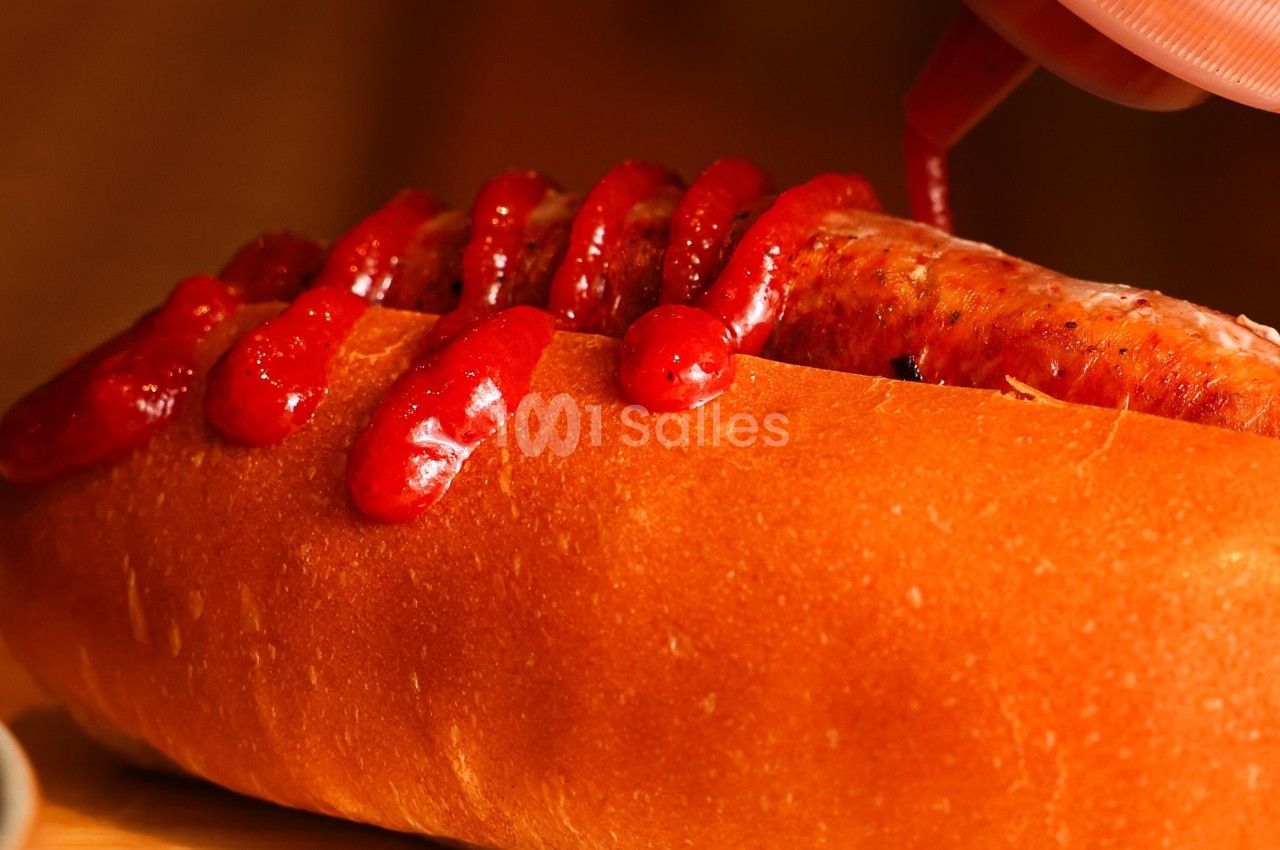 Saucisse grillée dans un pain allongé, garnie de ketchup en cours de versement.