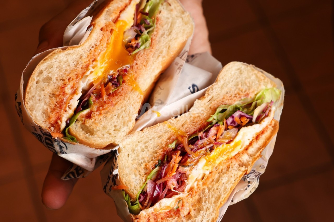 Sandwich coupé en deux, garni de légumes, fromage fondu et sauce, tenu dans une main sur un fond flou.