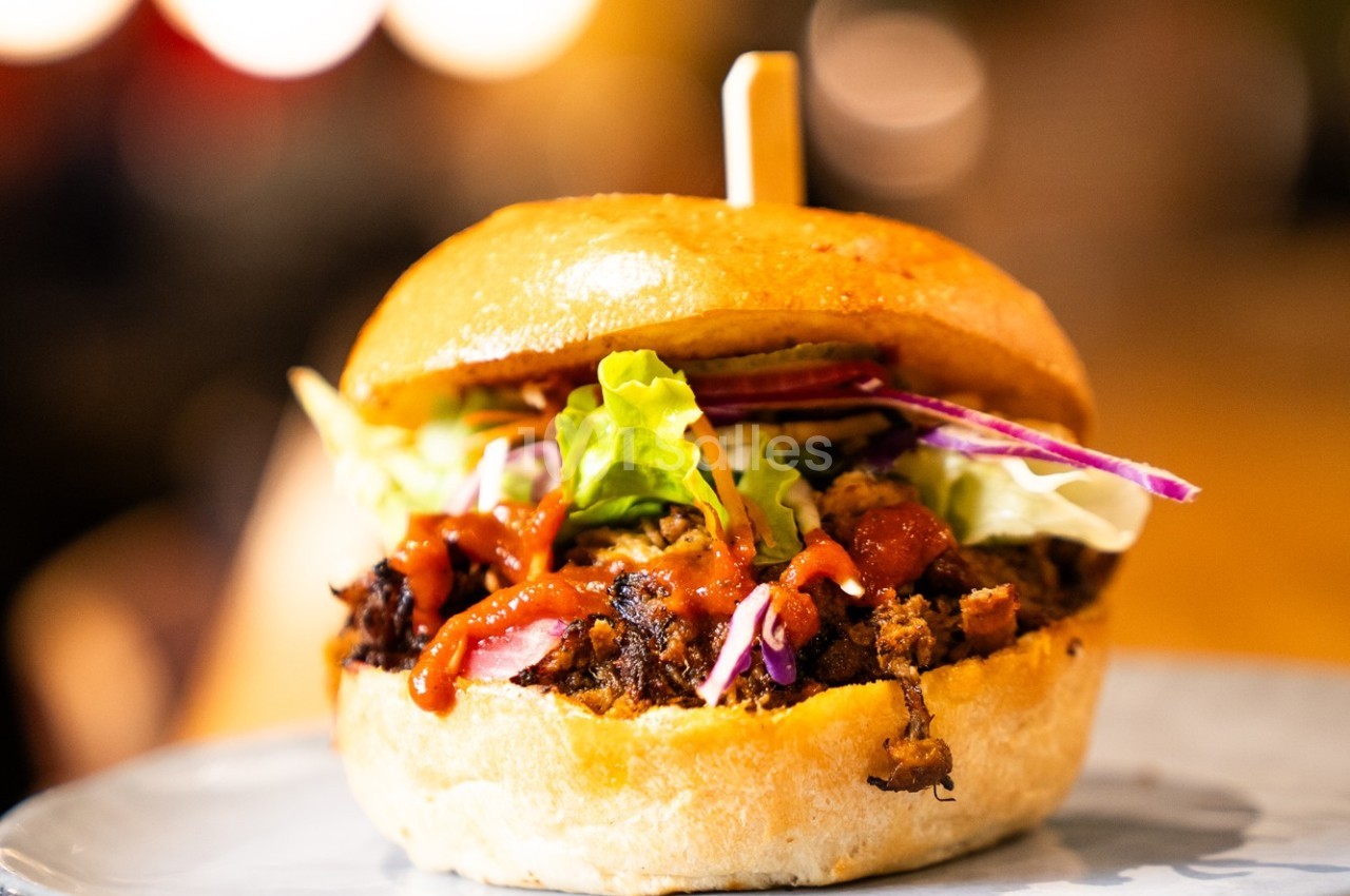 Burger garni de viande effilochée, salade, chou rouge et sauce, présenté sur une assiette avec un fond flou.