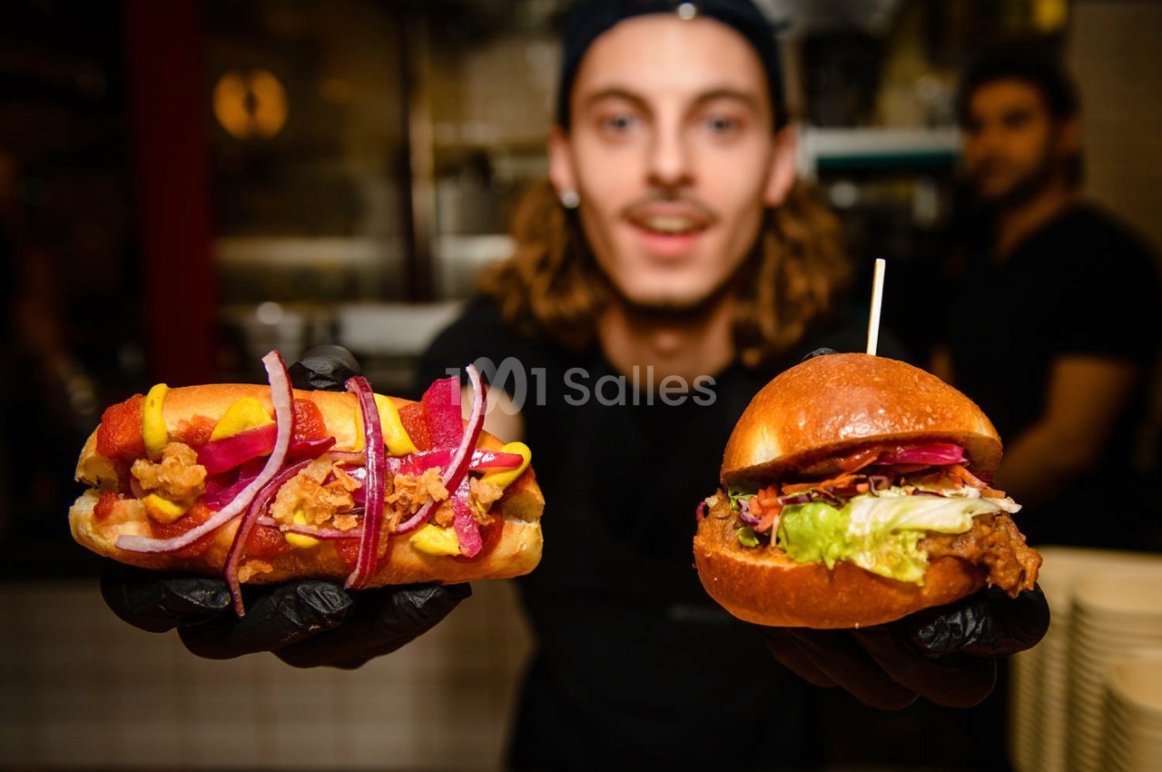 Un cuisinier présente un hot-dog garni et un burger avec des ingrédients frais dans un restaurant.