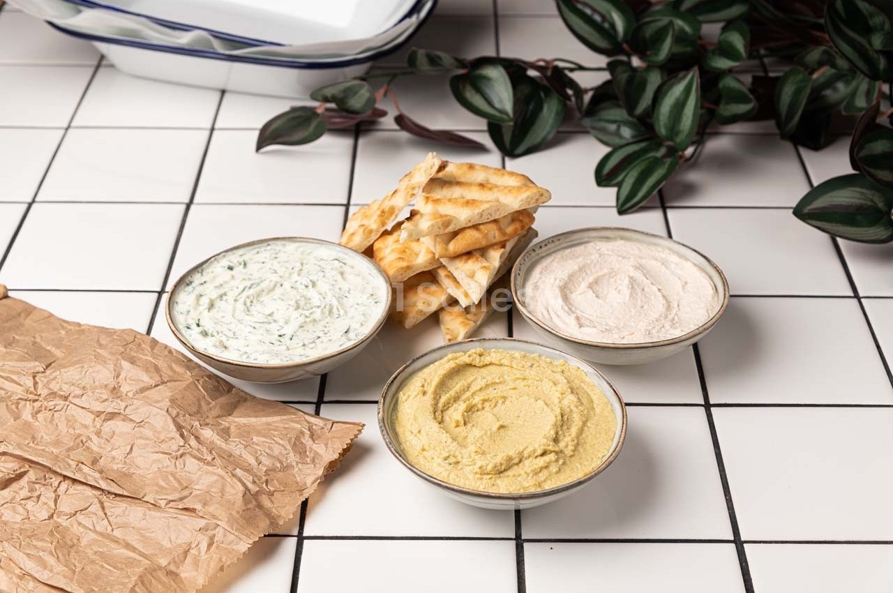 Trois bols de trempettes variées avec des crackers sur une table en carrelage blanc, décorée de feuilles vertes.