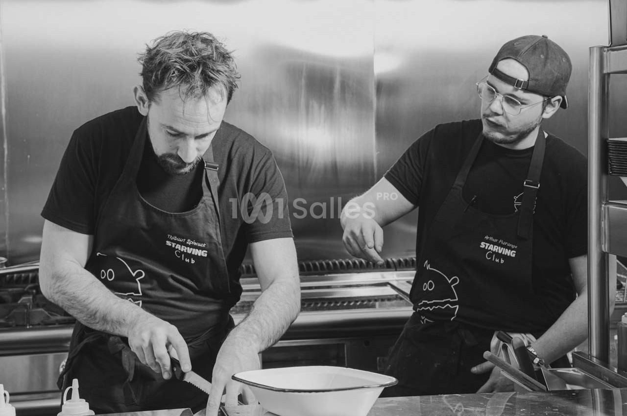 Deux cuisiniers en tabliers noirs préparent des ingrédients dans une cuisine professionnelle.