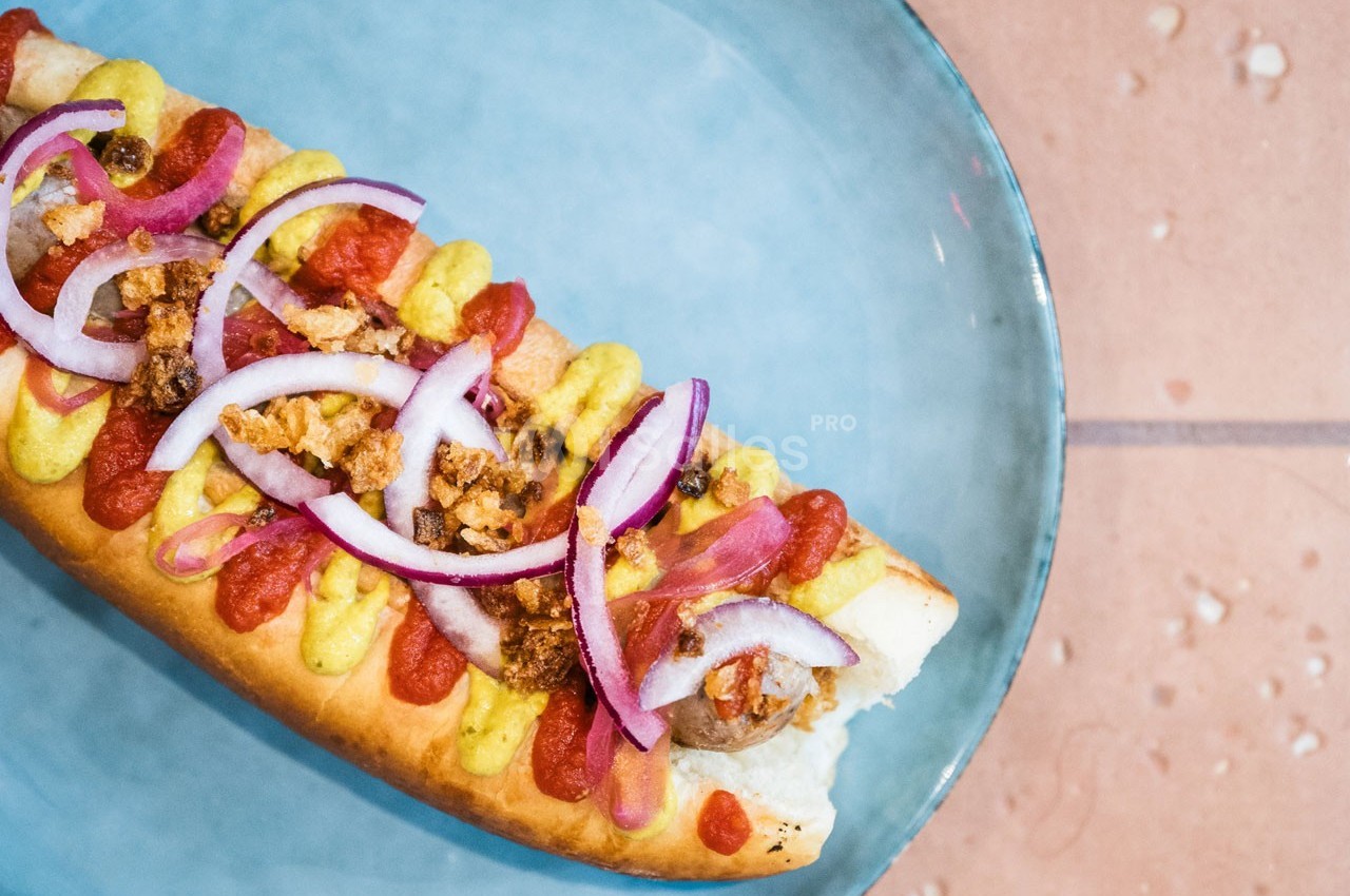 Hot-dog garni de moutarde, ketchup, oignons rouges, miettes croustillantes et saucisse, présenté sur une assiette bleue.