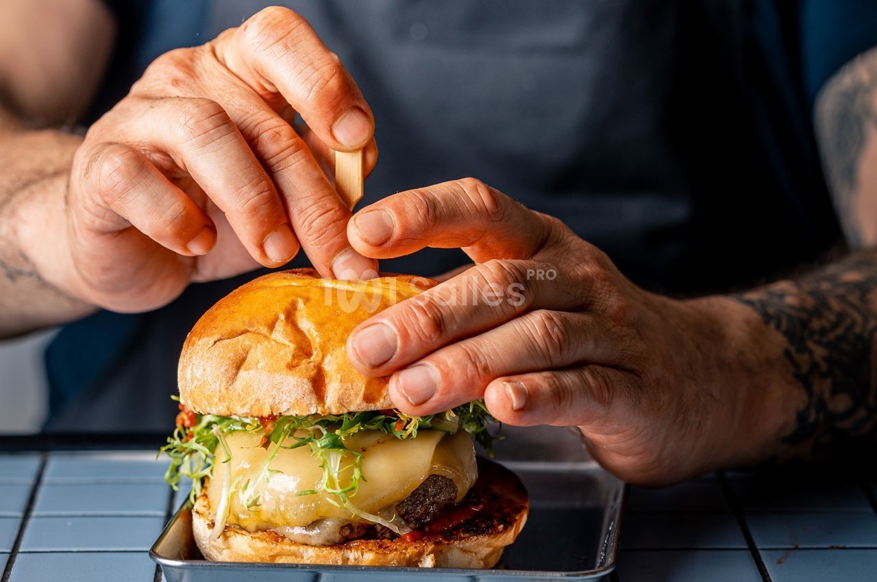 Un cuisinier assemble un burger garni de fromage fondu, de jeunes pousses et de sauce, posé sur un plateau métallique.