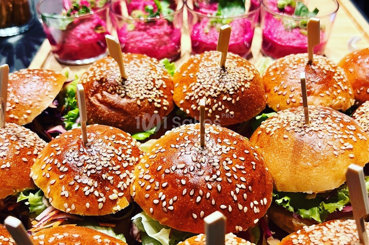 Mini burgers garnis de salade et de viande, avec des pains aux graines de sésame, accompagnés de verrines roses en arrière…