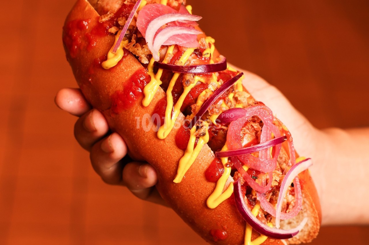 Hot-dog garni de moutarde, ketchup, oignons rouges et frits, tenu en main sur un fond flou orangé.