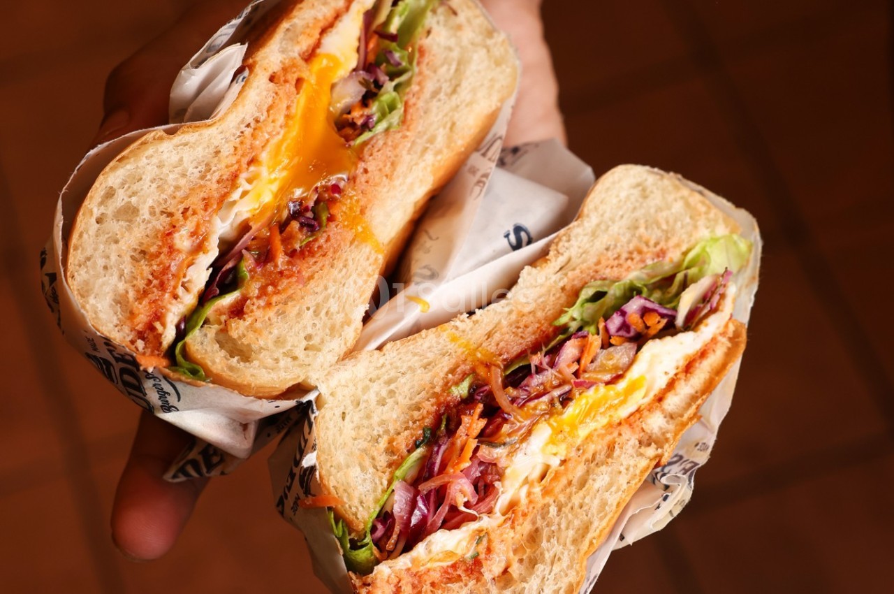 Sandwich coupé en deux, garni de légumes, fromage fondu et sauce, tenu dans une main sur un fond flou.