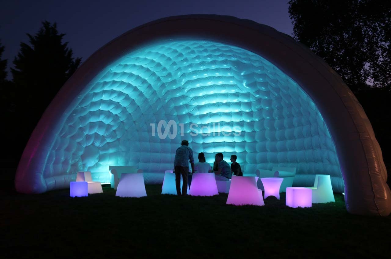 Structure gonflable éclairée en bleu et violet, avec des personnes assises autour de tables lumineuses dans l'obscurité.