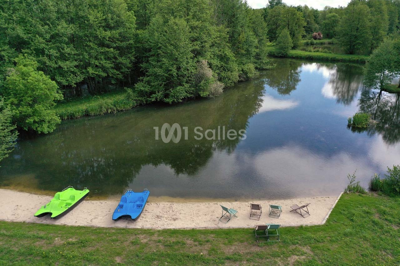 Vue d'un petit lac entouré de forêt avec deux pédalos colorés et des chaises disposées sur une plage de sable.