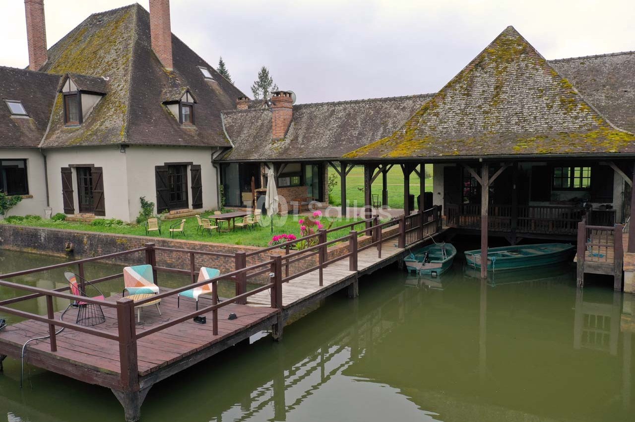 Ponton en bois avec chaises et table, donnant sur un plan d'eau, près d'une maison à toit en ardoise et jardin verdoyant.