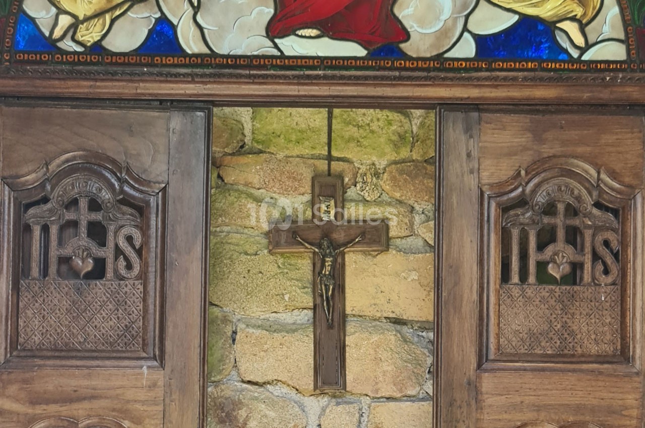 Croix en bois accrochée sur un mur en pierre, encadrée par des portes sculptées et un vitrail coloré au-dessus.