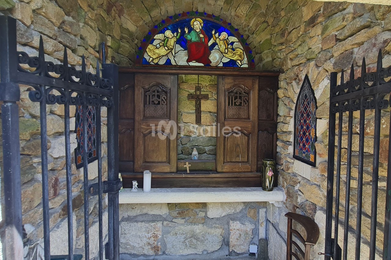 Autel en pierre avec croix en bois, vitrail coloré représentant une scène religieuse et grilles en fer forgé ouvertes.