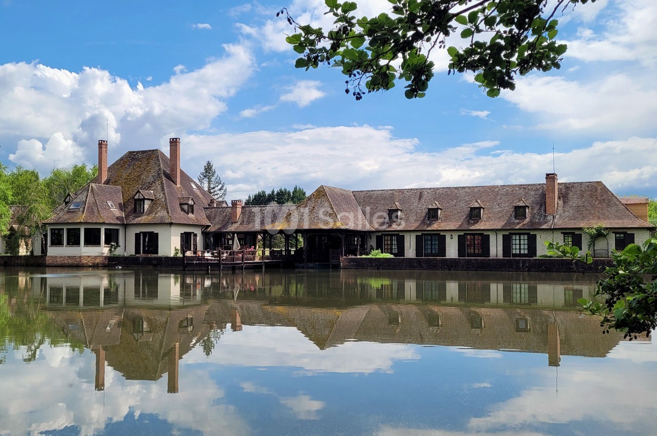 Grande maison au bord d'un étang calme, avec un ciel partiellement nuageux et des arbres en arrière-plan.