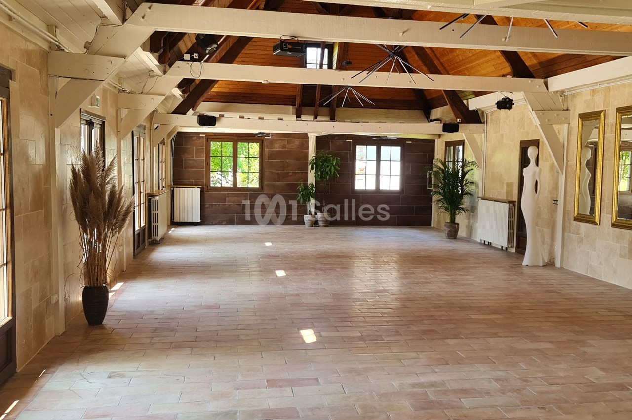 Salle lumineuse avec poutres apparentes, grandes fenêtres, plantes décoratives et statue blanche sur un sol en terre cuite.