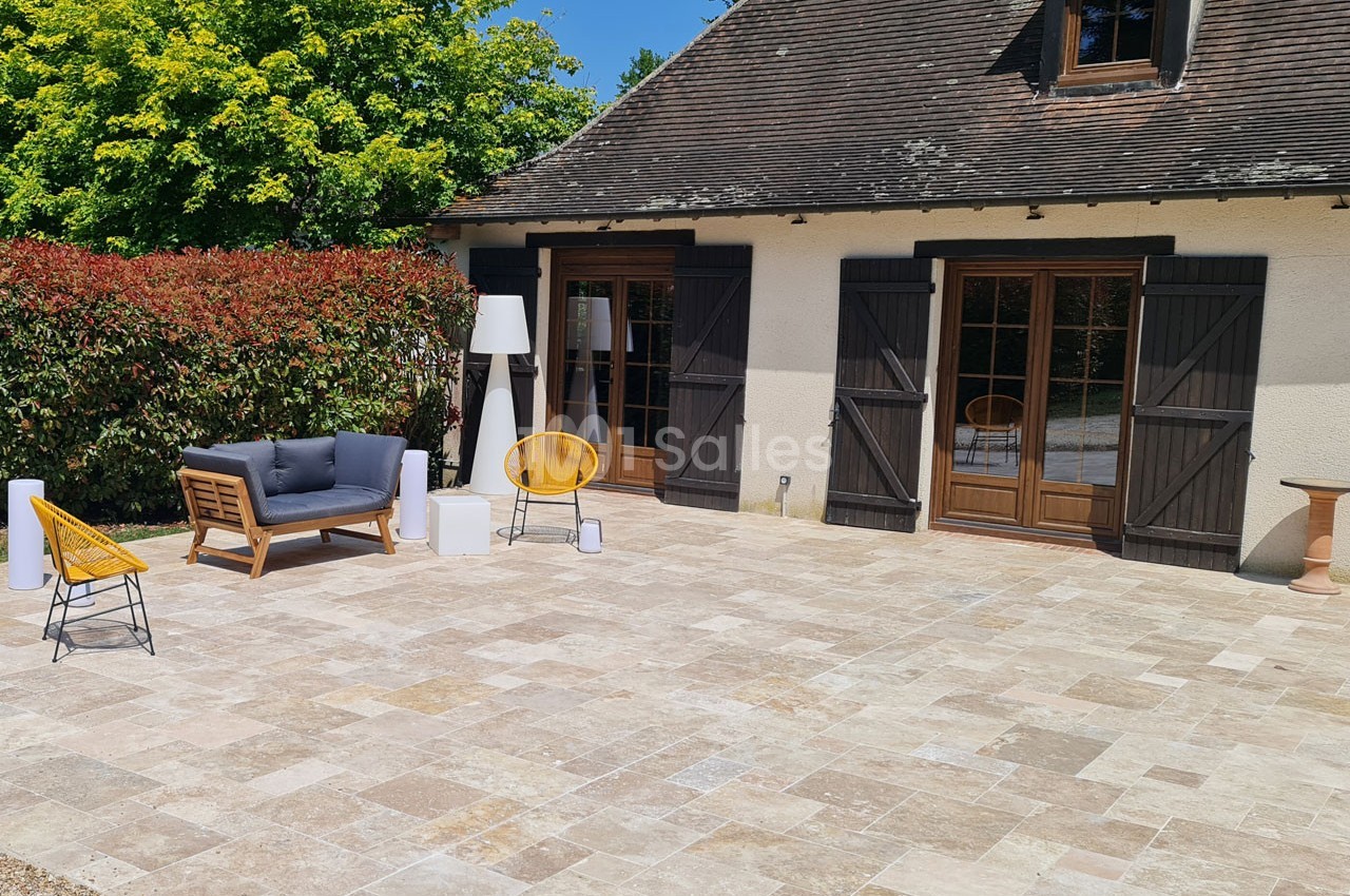 Terrasse en pierre avec mobilier de jardin moderne, devant une maison aux volets en bois et entourée de végétation.