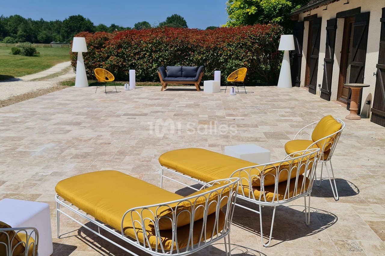 Terrasse en pierre avec mobilier de jardin jaune et blanc, entourée de haies rouges et d'un paysage verdoyant.