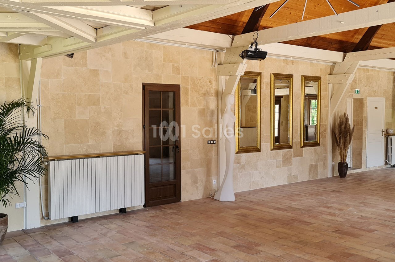 Salle lumineuse avec murs en pierre, sol en carrelage, poutres apparentes, miroirs dorés et plantes décoratives.
