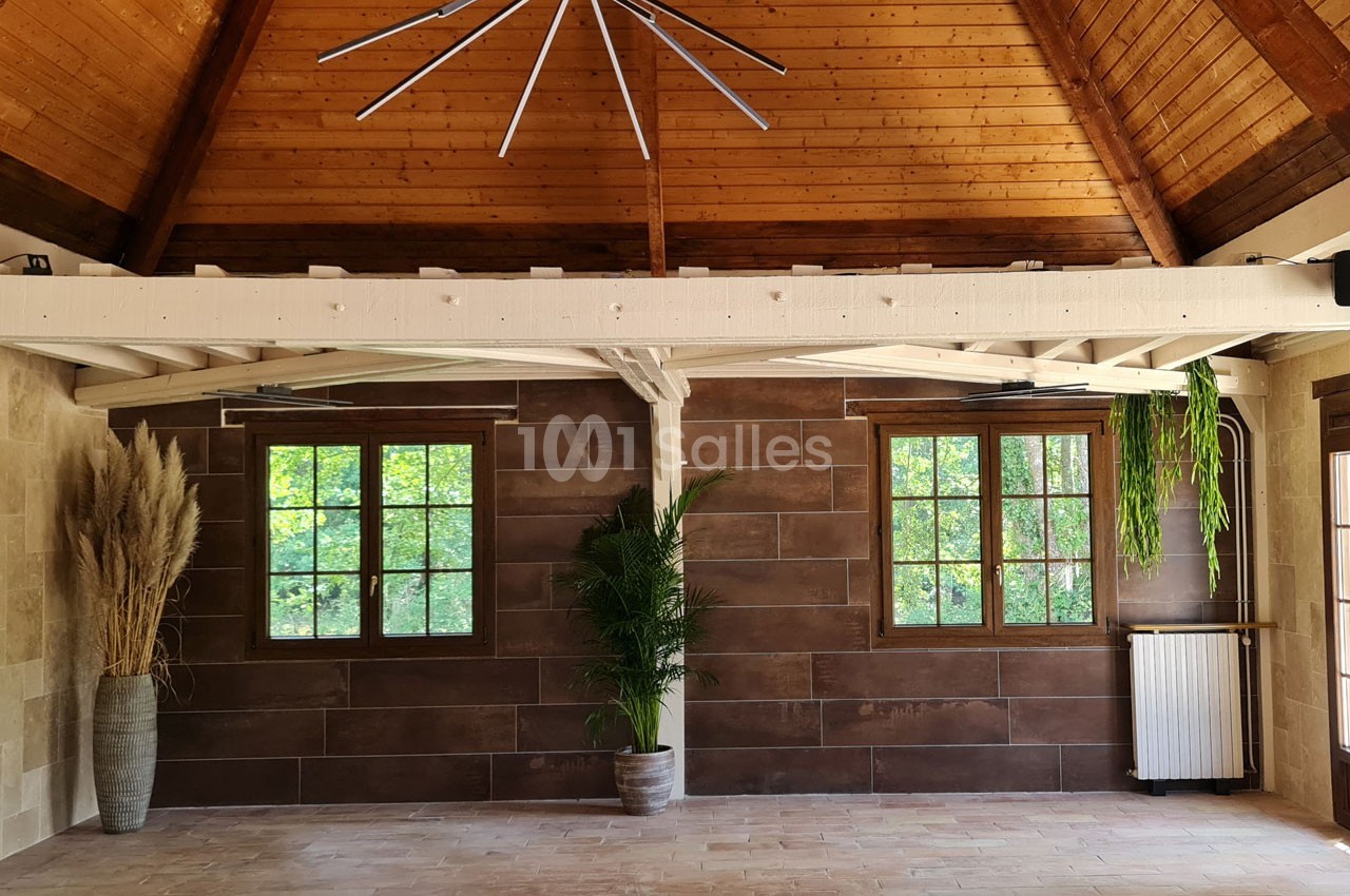 Salle lumineuse avec plafond en bois, murs en pierre et carrelage, deux fenêtres, plantes décoratives et radiateur.
