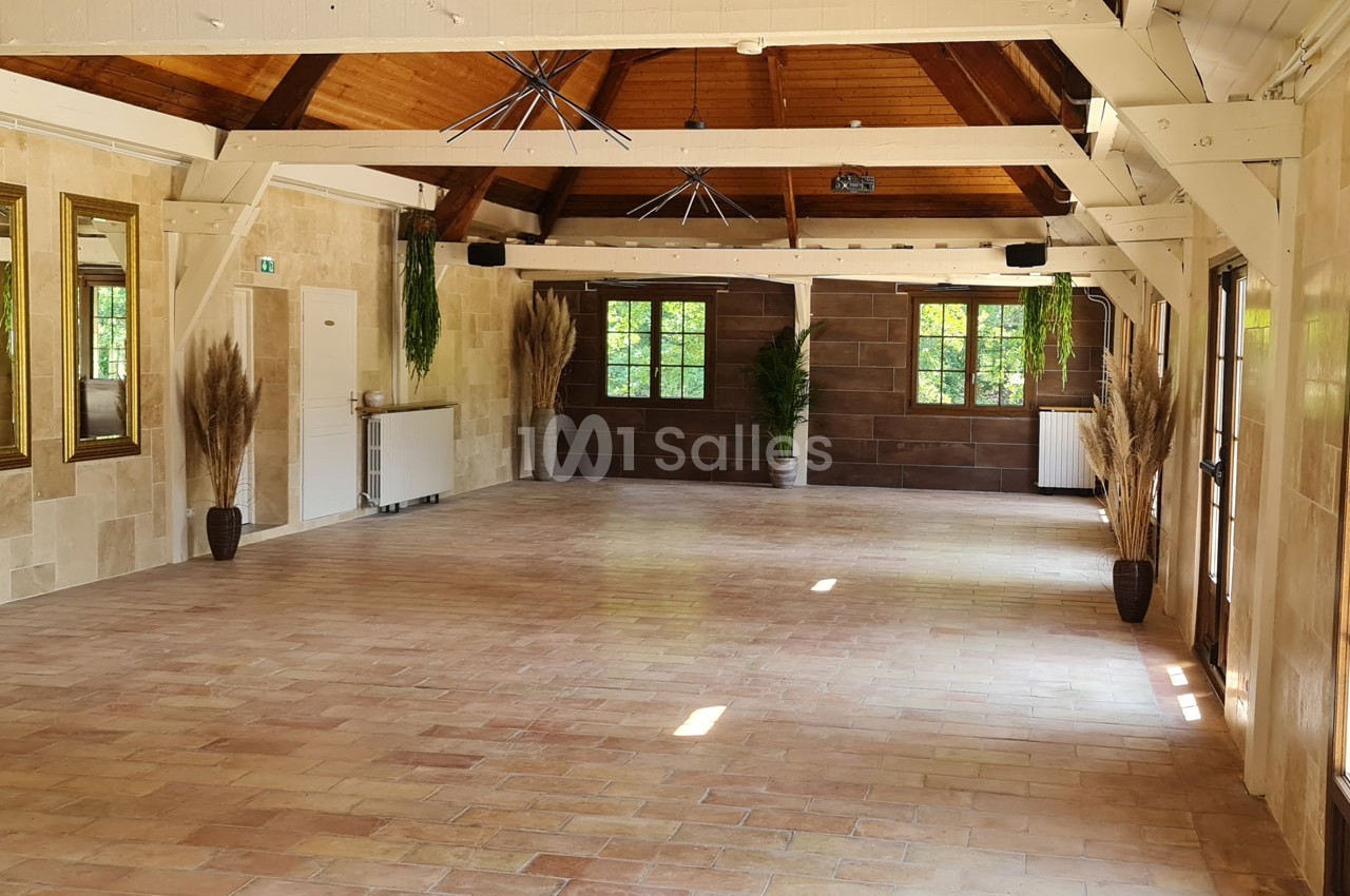 Salle lumineuse avec murs en pierre, poutres apparentes, grandes fenêtres, plantes décoratives et sol en carrelage.