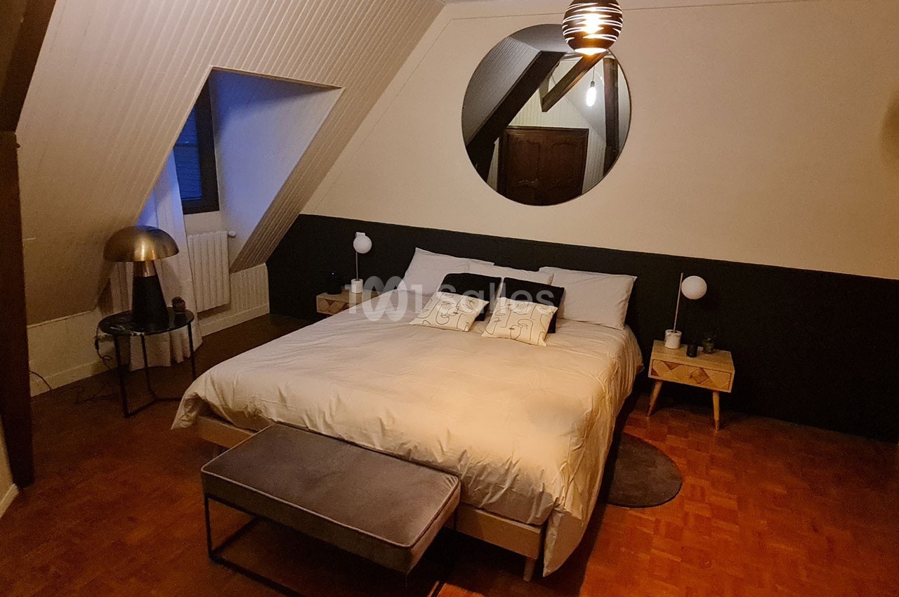 Chambre mansardée avec un lit double, tables de chevet, miroir rond au mur et éclairage tamisé.