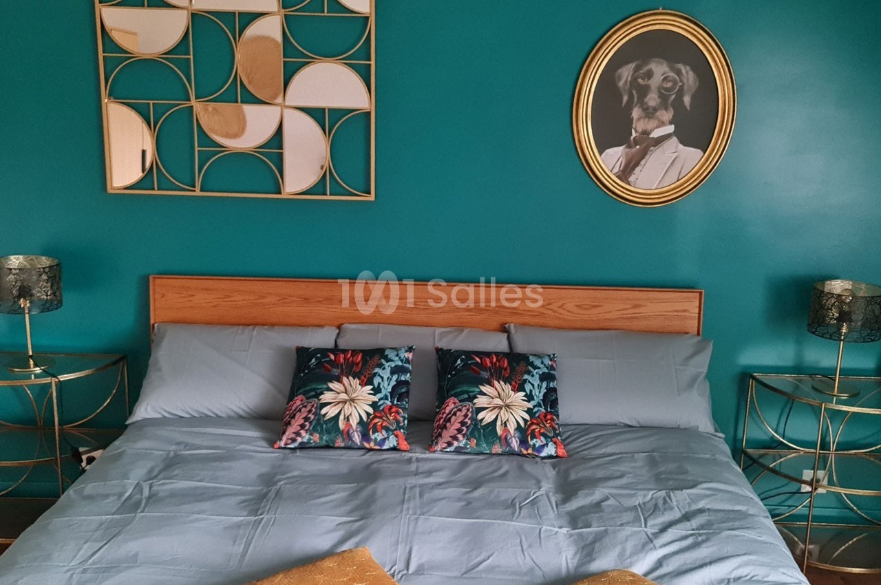 Lit double avec draps bleus, coussins à motifs floraux, mur turquoise décoré d'un tableau et d'un miroir géométrique.