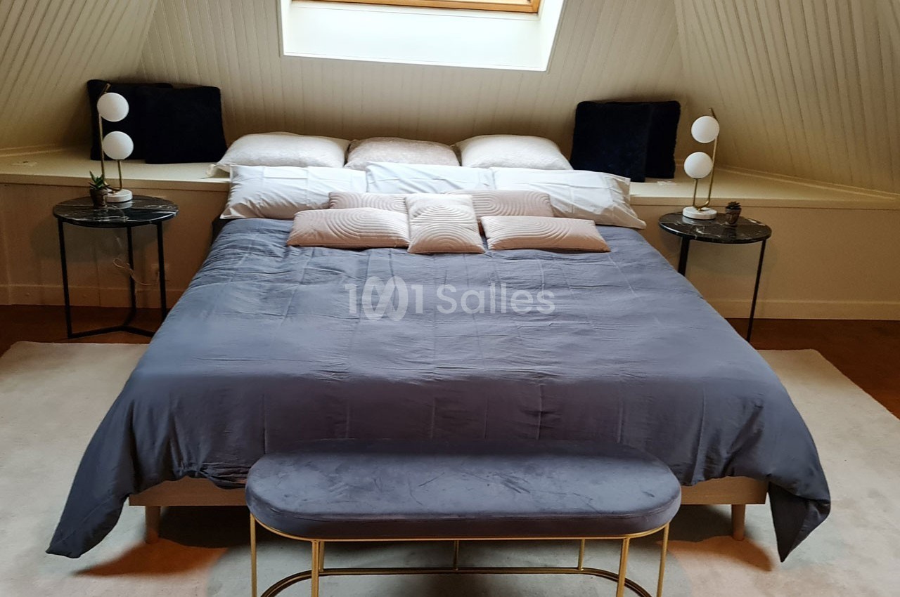 Lit double avec draps gris, coussins décoratifs et tables de chevet surmontées de lampes, dans une chambre mansardée.