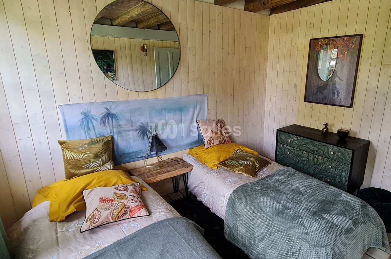 Chambre avec deux lits simples, murs en bois clair, décor tropical, miroir rond et commode verte.